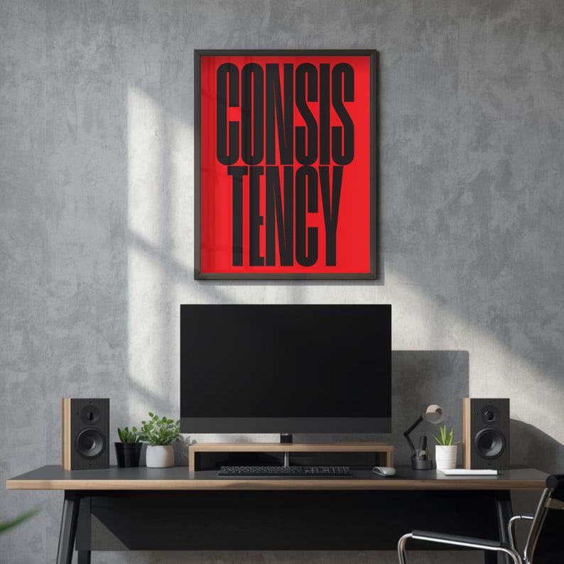 Op de afbeelding: Een ingelijste kunstprint met het woord "CONSISTENCY" in grote zwarte letters op een rode achtergrond. De print hangt boven een bureau met een tv, luidsprekers en planten. De kamer heeft een moderne, minimalistische uitstraling.