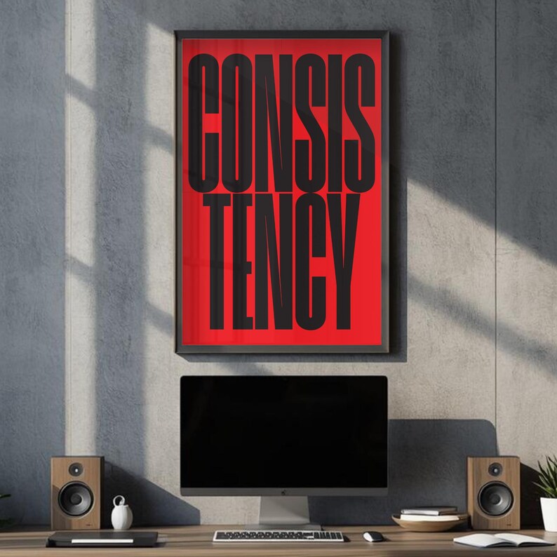 Op de afbeelding: Een ingelijste poster met het woord 'CONSISTENCY' in grote, zwarte, vette letters tegen een rode achtergrond. De poster hangt boven een computermonitor, luidsprekers en andere bureau-items. Het algemene thema is motivatie.