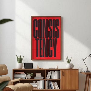 Op de afbeelding: Een ingelijste kunstdruk met het woord "CONSISTENCY" in vette, zwarte hoofdletters tegen een rode achtergrond. De print zit in een bruine houten lijst. Het kunstwerk wordt tentoongesteld aan een witte muur boven een houten kast.
