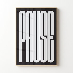May include: A rectangular wall art print in a light wood frame featuring the word PAUSE in large, vertical, white sans-serif capital letters against a solid black background. The typography is elongated and fills the entire height of the frame.