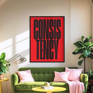Op de afbeelding: Een ingelijste kunstprint met het woord "CONSISTENCY" in zwarte letters op een rode achtergrond. Het kunstwerk hangt boven een groene bank met roze kussens en een gele salontafel. Moderne woonkamer.