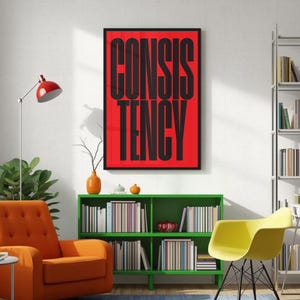 Op de afbeelding: Een ingelijste kunstdruk met het woord "CONSISTENCY" in vette zwarte letters op een rode achtergrond. De print hangt aan een witte muur boven een groene boekenkast. Een oranje fauteuil en een gele stoel staan ook in de kamer.