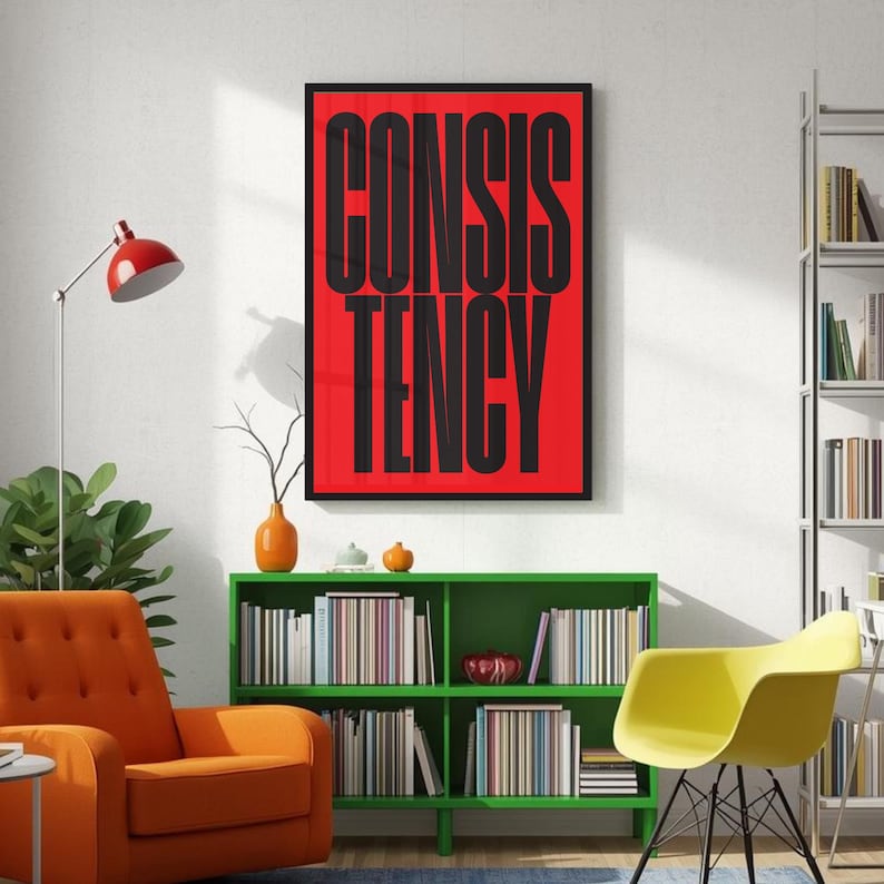 Op de afbeelding: Een ingelijste kunstdruk met het woord "CONSISTENCY" in vette zwarte letters op een rode achtergrond. De print hangt aan een witte muur boven een groene boekenkast. Een oranje fauteuil en een gele stoel staan ook in de kamer.