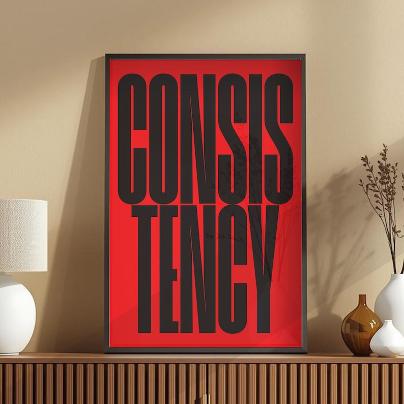 Op de afbeelding: Een ingelijste print met het woord "CONSISTENCY" in grote, zwarte hoofdletters op een rode achtergrond. Het kunstwerk wordt getoond op een houten kast, met een lamp en vazen op de achtergrond.