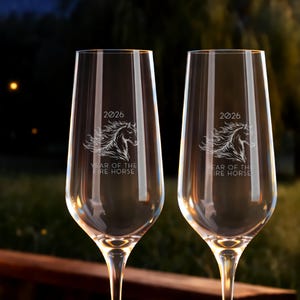 Op de afbeelding: Twee heldere champagneglazen met een paardenontwerp geëtst op het glas. De tekst "2026" en "Year of the Fire Horse" zijn ook op het glas geëtst. De glazen staan tegen een vage achtergrond.
