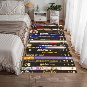 Puede incluir: Una alfombra rectangular con un diseño de cintas VHS apiladas. Se ven títulos de películas como "Halloween" y "Ghostbusters". La alfombra está colocada sobre un suelo de madera junto a una cama con ropa de cama blanca.