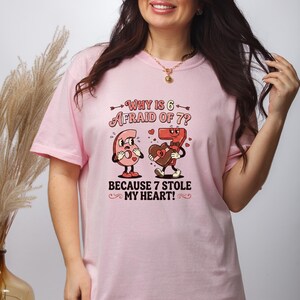 May include: Pink t-shirt with the text "Why is 6 afraid of 7? Because 7 stole my heart!" featuring cartoon numbers 6 and 7. Number 6 has a worried expression, while 7 holds a heart-shaped box.