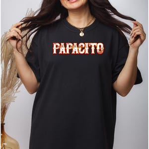 May include: Black t-shirt with the word "PAPACITO" in a red and white decorative font. The shirt is worn by a person with long dark hair and a gold necklace.