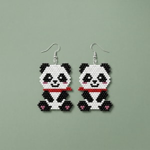 May include: A pair of panda bear earrings. Each earring is a black and white panda design with a red scarf and pink details. The earrings have silver-coloured fish hook ear wires. The background is a muted green.