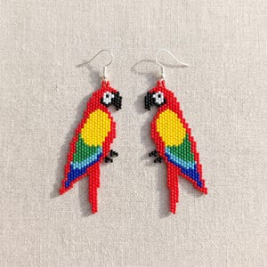 May include: A pair of beaded parrot earrings. Each earring features a red body, yellow chest, and a rainbow of colors on the wings. The parrot's head is black with a red spot. The earrings hang from silver hooks.