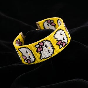 May include: A yellow beaded bracelet featuring a repeating pattern of a white cat face with a pink bow. The bracelet has a gold clasp and is set against a black background. The bracelet is a fun and colorful accessory.
