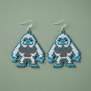 May include: A pair of handmade beaded earrings in the shape of a yeti. The earrings feature a blue, white, and gray color scheme. The yeti design has a triangular shape with silver-colored ear wires. The background is a solid green color.