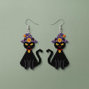 Peut inclure: Une paire de boucles d'oreilles en perles représentant un chat noir. Chaque chat porte un chapeau de sorcière violet avec des détails orange et jaunes, un collier rouge et a des yeux jaunes. Les boucles sont faites de perles et de crochets argentés.
