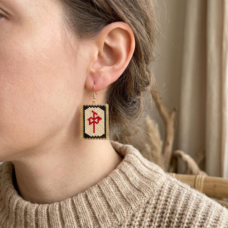 May include: Gold-tone drop earrings featuring a rectangular design with a beaded pattern. The earrings have a cream-colored center with a red symbol, framed by black and gold beads. The earrings are worn on a person's ear.