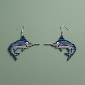 Op de afbeelding: Een paar kralen oorbellen met een zwaardvis ontwerp. De oorbellen zijn voornamelijk blauw, met grijze en witte accenten, en zilverkleurige oorhaken. De zwaardvissen hebben een lange, puntige snuit en een gedetailleerd, getextureerd uiterlijk.