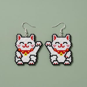 Puede incluir: Un par de pendientes de gato blanco con contornos negros, detalles rojos y un collar rojo y amarillo. Los pendientes tienen la forma de un gato saludando, con ganchos plateados. El fondo es verde liso y apagado.