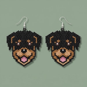 May include: A pair of beaded drop earrings shaped like a Rottweiler dog head. The design features black, tan, and brown beads forming the facial features, with a small pink bead representing the tongue and silver-toned metal earring hooks.