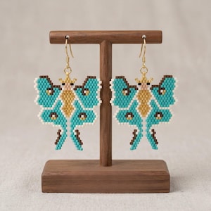 Puede incluir: Un par de pendientes de mariposa con cuentas en turquesa, marrón, dorado y blanco, exhibidos en un expositor de joyas de madera. Los pendientes presentan un intrincado trabajo de cuentas, con ganchos dorados.