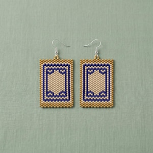 Könnte beinhalten: Ein Paar rechteckige Perlenohrringe. Die Ohrringe haben einen goldenen Rand, eine cremefarbene Mitte und ein marineblaues geometrisches Design. Die Ohrringe hängen an silberfarbenen Haken.
