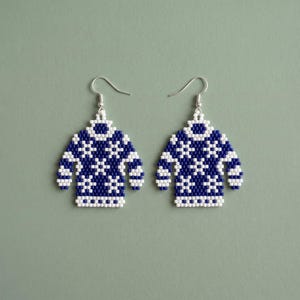 Op de afbeelding: Een paar hangende oorbellen in de vorm van miniatuurtruien, gemaakt van kleine donkerblauwe en witte kralen. Het truiontwerp heeft een herhalend geometrisch sneeuwvlokpatroon aan de voorzijde, afgewerkt met zilverkleurige metalen oorhaken.