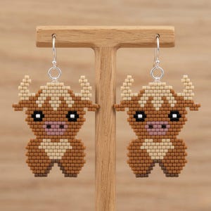 Peut inclure: Boucles d'oreilles faites à la main en forme de vaches des Highlands. Les boucles d'oreilles sont marron, beige et rose, avec des crochets d'oreilles argentés. Les vaches ont un design détaillé et pixélisé, avec des cornes et un visage mignon.