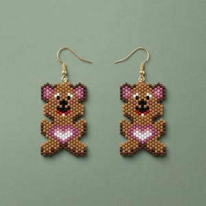May include: A pair of dangle earrings featuring a beaded teddy bear design. The bears are crafted from brown, tan, pink, white, and black beads, with a pink and white heart on the chest. Each earring hangs from a gold-toned metal hook wire.