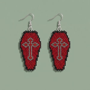 May include: A pair of drop earrings featuring a coffin-shaped design crafted from small beads. The earrings have a red background with a central grey cross pattern, all outlined in black beads, and are attached to silver-toned metal hooks.