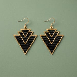 May include: A pair of geometric earrings with a black and gold design. The earrings are triangular, with a layered chevron pattern. The edges and the chevron lines are gold, and the rest of the earring is black. The earrings have gold-colored ear wires.