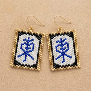 Op de afbeelding: Een paar rechthoekige, met kralen bezette oorbellen. Elke oorbel heeft een witte achtergrond met een blauw symbool, omlijst door zwarte en gouden kralen. De oorbellen hebben goudkleurige oorhaken.