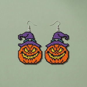May include: A pair of beaded dangle earrings featuring a jack-o'-lantern pumpkin design with a purple witch hat. The pumpkin is orange with yellow eyes and a yellow mouth, while the hat has a teal band. Silver-toned metal earring hooks.