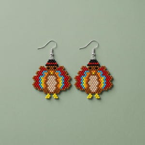 May include: A pair of beaded dangle earrings shaped like turkeys. Each earring features a central tan body, a brown head with a black hat, and a fan-shaped tail made of red, yellow, and blue beads. The earrings hang from silver-toned metal hooks.