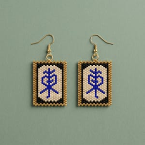 May include: A pair of beaded earrings with a rectangular design. The earrings feature a gold border, a black background, and a cream-colored center with a blue symbol. The earrings hang from gold-colored ear wires.