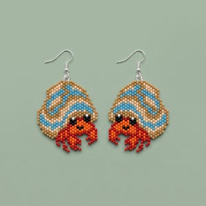 May include: A pair of dangle earrings crafted from small beads, shaped like hermit crabs. The shells feature horizontal stripes in light blue, tan, and white, while the crab bodies are bright orange with black eyes and silver-toned metal hooks.