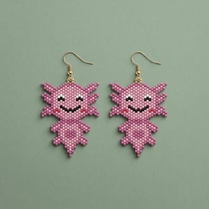 May include: A pair of pink axolotl earrings with gold-colored hooks. The earrings are made of small beads, with a smiling face and a frilly design. The background is a solid, muted green.