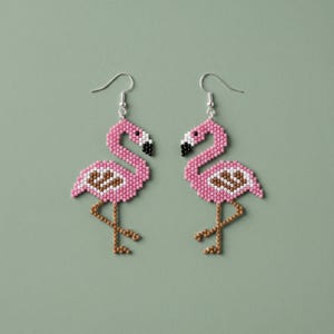 May include: A pair of flamingo earrings. Each earring is crafted from small pink beads, with brown and white accents on the body and a black beak. The earrings hang from silver-colored hooks, set against a muted green background.