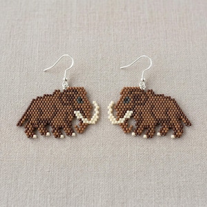 May include: A pair of handmade earrings featuring a pixelated design of a woolly mammoth. The earrings are made with small brown beads, with off-white tusks and feet, and blue eyes. The earrings hang from silver-colored hooks.