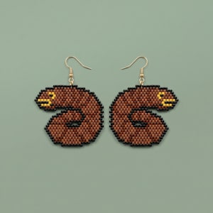 May include: A pair of handmade beaded earrings in a unique, abstract design. The earrings feature a brown, comma-shaped pattern with black and yellow accents, suspended from gold-colored ear wires. The earrings are set against a muted green background.