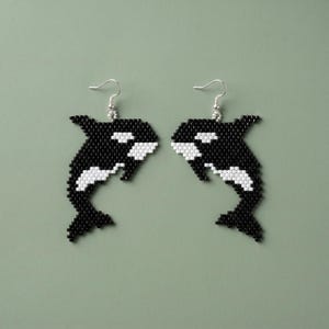 May include: A pair of beaded earrings featuring a black and white orca design. The earrings have a silver hook and are set against a muted green background. The orcas are detailed with white accents.