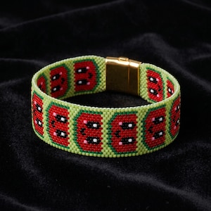 Könnte beinhalten: Ein Perlenarmband mit einem sich wiederholenden Muster aus roten Wassermelonenscheiben mit schwarzen Kernen und weißen Akzenten auf hellgrünem Hintergrund. Das Armband wird mit einem rechteckigen, goldfarbenen Metall-Magnetverschluss geschlossen.