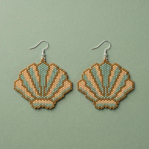 May include: A pair of beaded seashell earrings. The earrings feature a fan-shaped design with alternating stripes of light blue, tan, and brown beads. The earrings have silver-colored ear wires and are set against a muted green background.