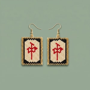 May include: A pair of gold-coloured drop earrings. Each earring has a rectangular pendant with a black border, a cream-coloured centre with a red symbol, and a gold beaded frame. The earrings hang from gold-coloured fish hook ear wires.