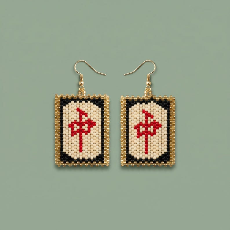 May include: A pair of gold-toned drop earrings featuring rectangular pendants. Each pendant has a black border, a cream-colored center with a red symbol, and a gold beaded frame. The earrings hang from gold-colored fish hook ear wires.
