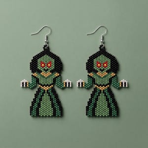 May include: A pair of beaded dangle earrings featuring a pixelated creature design. The figure has a black head, bright red eyes, and a green body with gold accents. The earrings are constructed from small, uniform beads and hang from silver-toned hooks.