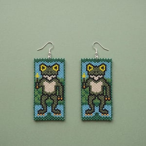 May include: A pair of rectangular beaded earrings featuring a pixelated frog design. The frog is dark green with a white chest, yellow eyes, and holds a small yellow object. The background consists of blue and green square beads with silver hooks.