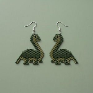 May include: A pair of handmade dinosaur earrings. Each earring features a beaded design in shades of green and tan, shaped like a Brontosaurus. The earrings have silver-colored fish hook ear wires. The background is a solid, muted green.