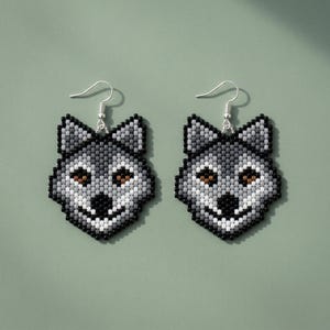 Op de afbeelding: Een paar kralen oorbellen met een wolf. Elke oorbel heeft een wolfskopontwerp in grijstinten, zwart en wit, met bruine ogen. De oorbellen hebben zilverkleurige oorhaken en een gedetailleerd, pixelachtig uiterlijk.