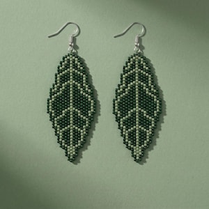 May include: A pair of dangle earrings featuring a leaf-shaped design crafted from small dark green and light green seed beads. The earrings hang from silver-toned metal hooks and display a symmetrical pattern of a central vein with side veins.