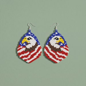 May include: A pair of beaded earrings in a teardrop shape, featuring a patriotic design. The earrings depict an American bald eagle with a blue border, a white and red striped pattern, and a brown eagle body. The earrings hang from silver hooks.
