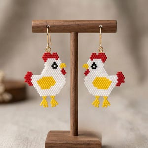 Peut inclure: Une paire de boucles d'oreilles poulet en perles. Chaque boucle d'oreille présente un corps blanc, un ventre jaune, une crête et une queue rouges, et des pieds jaunes. Les boucles d'oreilles sont présentées sur un présentoir en bois avec des crochets dorés.