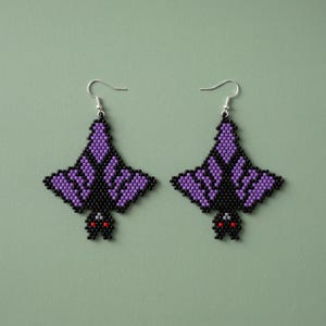 Pode incluir: Um par de brincos de morcego com contas. Os brincos apresentam um design de morcego preto com asas roxas e olhos vermelhos. Penduram-se em ganchos de orelha prateados. O fundo é verde-acinzentado.